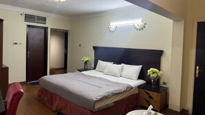 A hotel room with a bed, bedside tables, a nightstand, a desk, a chair, a small round table, a vase with flowers, and a door.