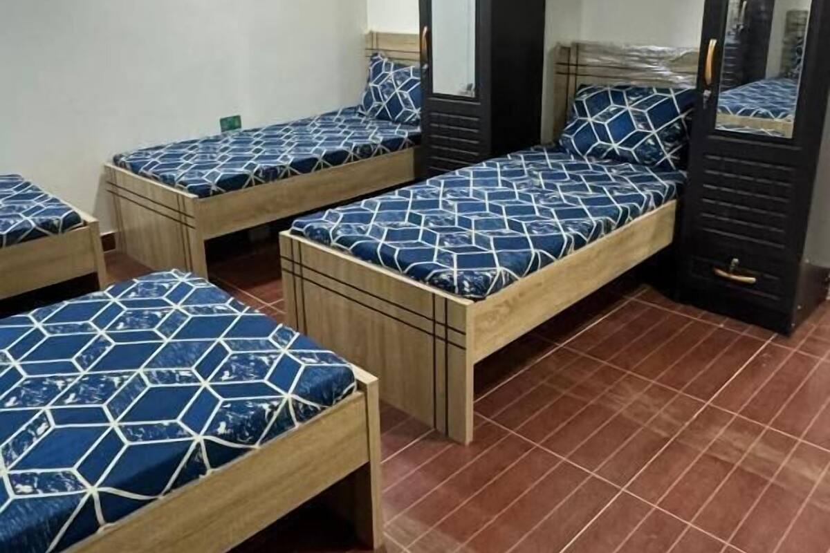 A dormitory room with four wooden beds, each with a blue and white patterned bedspread. There are two black wardrobes and a mirror on the wall.