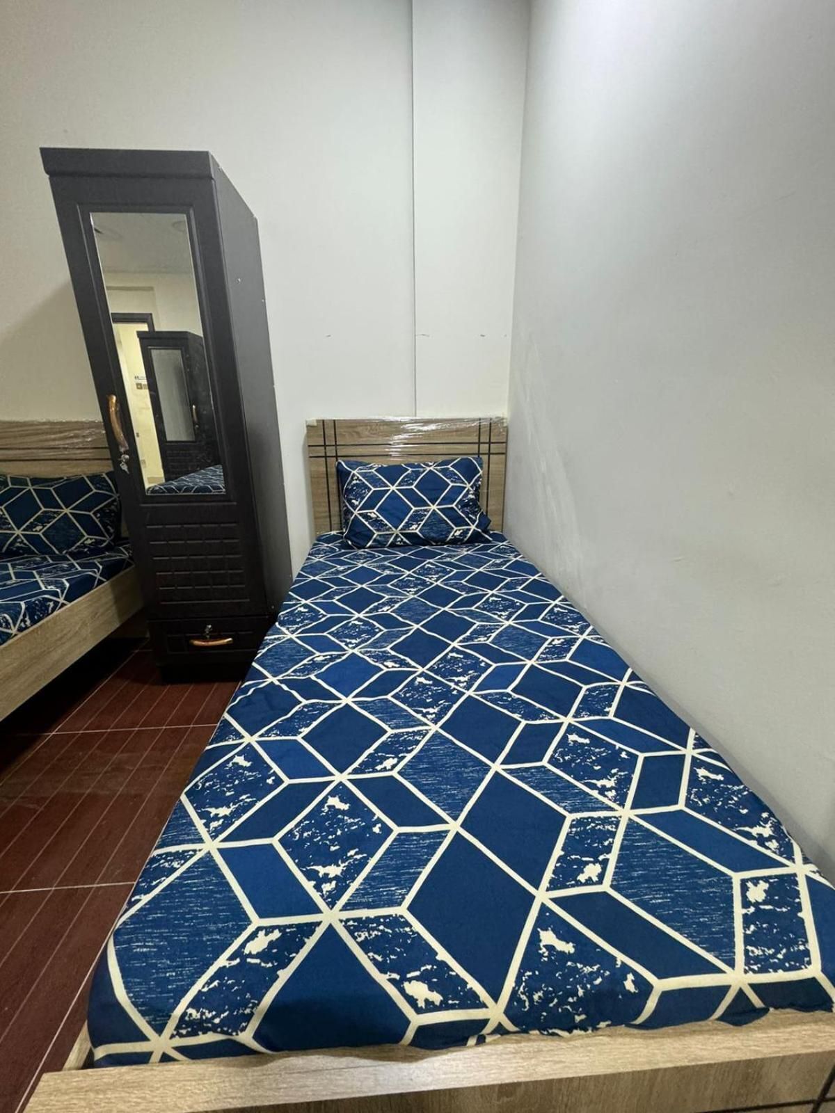 Une petite pièce étroite, avec un lit simple recouvert d’une couvre-lit aux motifs géométriques bleus et blancs.
