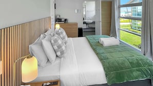A hotel room with a large bed, a bedside table with a lamp, a mirror, a window with curtains, and a view of the outside.