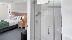 A modern hotel room with a glass-enclosed shower, a neatly made bed, and a bedside table with a lamp.