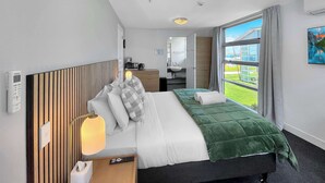 A hotel room with a bed, bedside table, lamp, and a view of a building through a window.