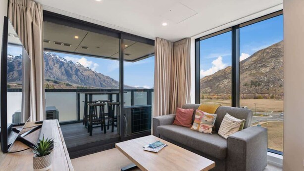 A modern hotel room with a balcony, a sofa, a coffee table, and a desk with a laptop. - Modern apartment with fantastic mountain views! (Queenstown)