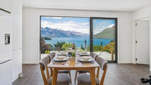 A dining area with a table set for four, a view of a lake and mountains through a glass door, and a modern kitchen to the left.