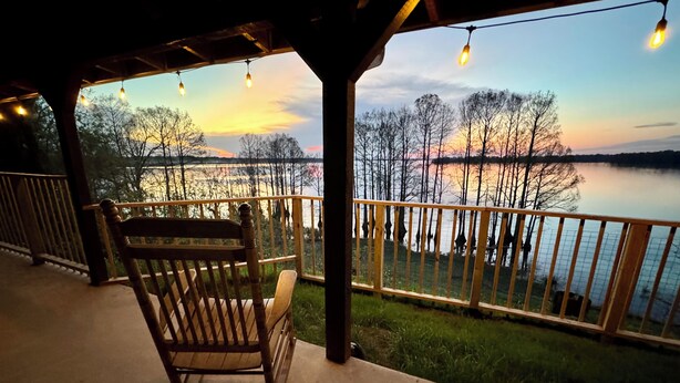 Una mecedora de madera en un porche cubierto con vista a un lago y un atardecer.