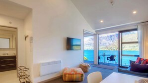 Un soggiorno moderno con una grande finestra che offre vista su montagne e un lago. Si vedono un divano, un tavolo da pranzo e una TV fissata al muro.