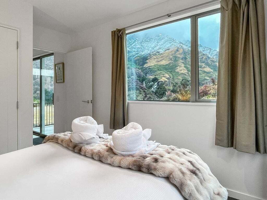 A hotel room with a bed, a window with curtains, a view of mountains, and a door leading to a balcony.
