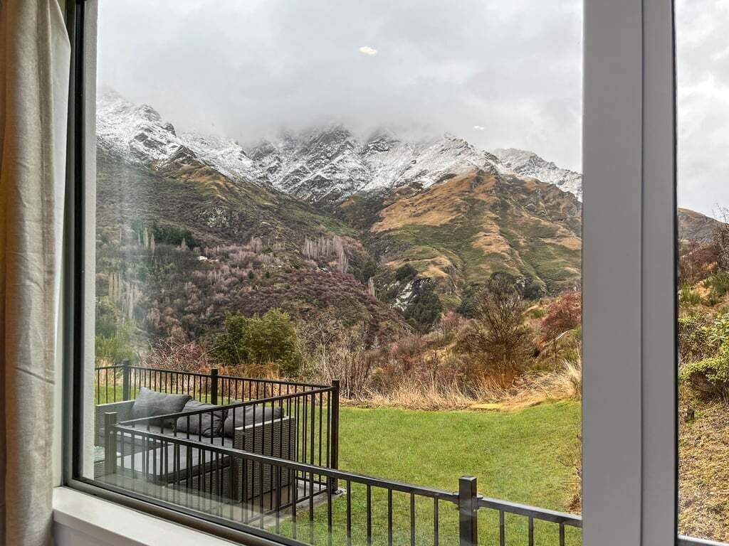 A view from a window with a balcony, overlooking snow-capped mountains and a grassy landscape.