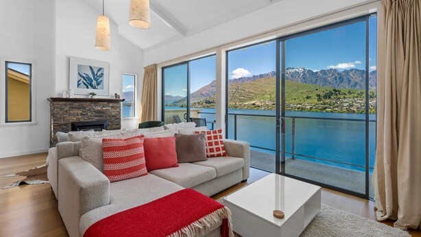 A modern living room with a large sofa, a coffee table, and a fireplace. There is a large window with a view of mountains and a lake.