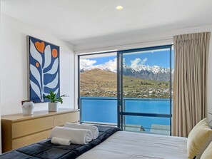 A hotel room with a large window offering a mountain view, a bed with neatly folded towels, a bedside table with a vase and a plant, and a modern abstract painting on the wall.