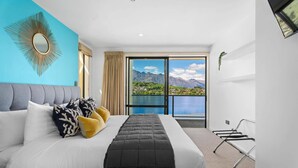 A modern hotel room with a large bed, a view of mountains and a lake, and a balcony with metal chairs.