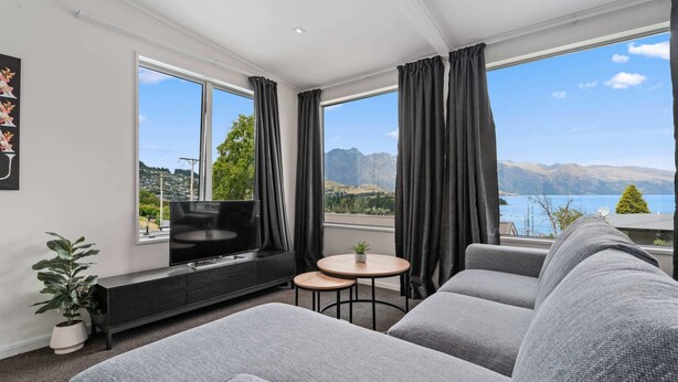 Un salon moderne avec un canapé gris, un téléviseur, une petite table sur laquelle est posée une plante, et de grandes fenêtres offrant une vue sur la montagne et le lac.