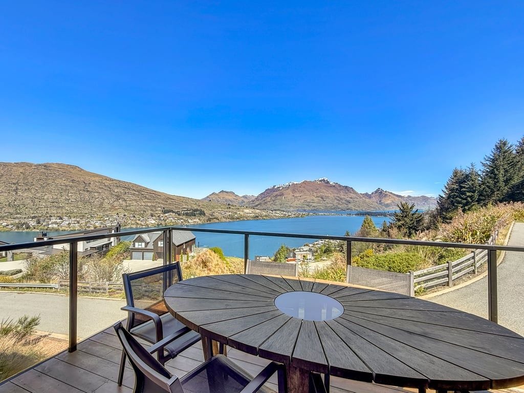 Een houten terras met tafel en stoelen met uitzicht op een meer en bergen.