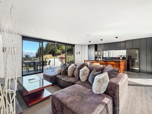 A modern living room with a large sectional sofa, a glass dining table, and a kitchen in the background. - Mountain Lake Vista 3 Bedroom (Queenstown)