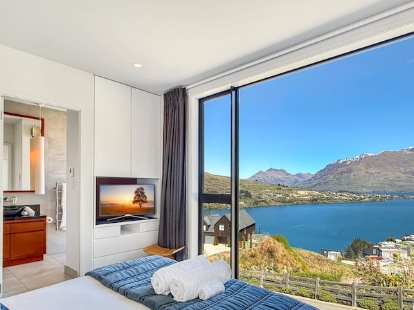 A modern bedroom with a large window, a bed, a TV, and a view of a lake and mountains.