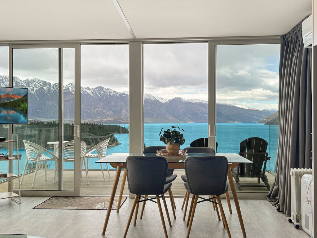 A dining area with a table and chairs, a view of mountains and water, and a glass door leading to an outdoor deck.