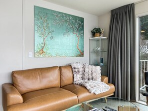 A living room with a brown leather sofa, a glass coffee table, a painting of a tree, a glass cabinet, and a grey curtain.