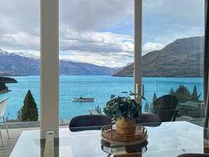 A room with a glass table, chairs, and a view of a lake with mountains in the background.