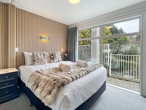 A bedroom with a large bed, a wooden wall panel, a nightstand, a lamp, and a view of a balcony.