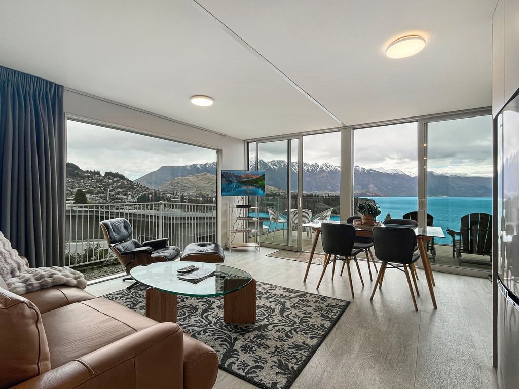 A modern living room with a glass table, leather sofa, and a view of mountains and a body of water.