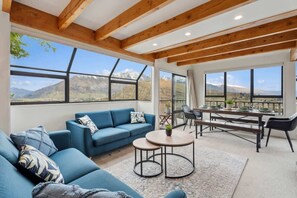 A spacious living area with a blue sofa, a round coffee table, and a dining table set for four. Large windows offer a view of mountains.