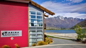 A red building with a sign reading "Marina Apartments" and a view of mountains and water in the background. - Marina 303A-Studio (Queenstown)