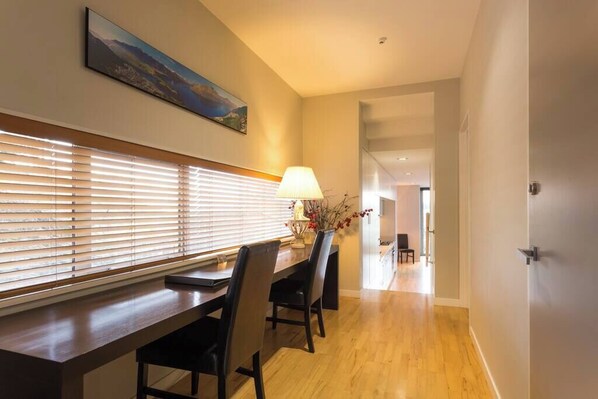 A dining area with a wooden table, chairs, and a lamp. There is a painting on the wall and a window with blinds.