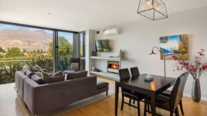 Een moderne woonkamer met een groot raam, een open haard, een eethoek en een aan de muur bevestigde televisie.