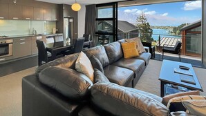 Una sala de estar moderna con sofá de cuero, comedor y cocina. Hay un ventanal que ofrece vistas al mar y a las montañas.