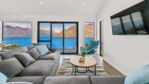 A modern living room with a large sofa, a round coffee table, and a view of a lake and mountains through large windows. - Lakeview Paradise On Wynyard (Queenstown)