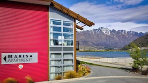 Un immeuble rouge avec une enseigne indiquant « Marina Apartments », et en arrière-plan, une vue sur des montagnes et l’eau.
