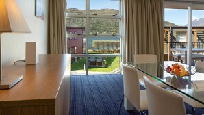 Une chambre d’hôtel dotée d’une grande fenêtre, d’un bureau en bois, d’une table en verre avec des chaises, et offrant une vue sur l’extérieur.