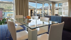 Une chambre d’hôtel moderne avec une table à manger en verre, des chaises blanches, un canapé et de grandes fenêtres offrant une vue sur les montagnes et un lac.