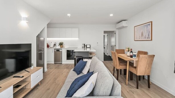 Una sala moderna con un televisor de pantalla plana, un sofá gris con cojines azules, una mesa de comedor de madera con sillas, y una cocina al fondo.