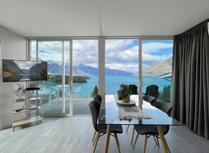 A modern room with a large window, a dining table, chairs, a TV, and a plant. - Spectacular VIEWS, 2 Bedrooms & BIG DECK! (Queenstown)
