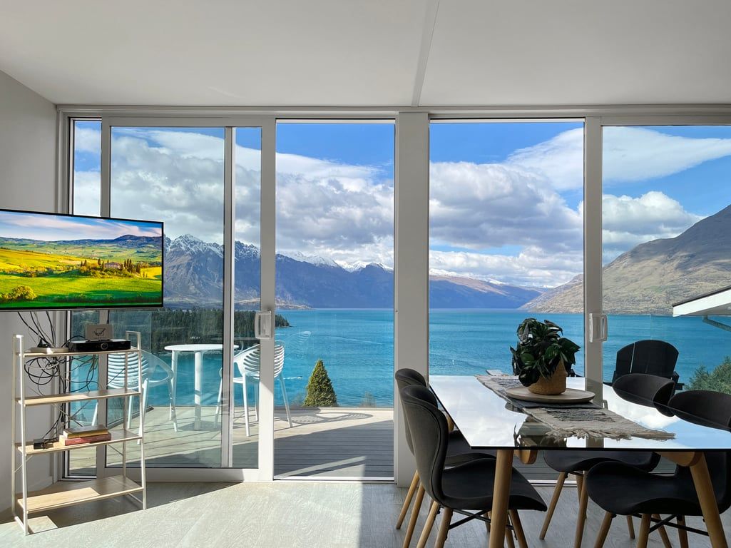 A modern room with a large window, a flat-screen TV, a dining area with chairs, and a small table.