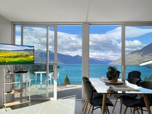 A modern room with a large window, a flat-screen TV, a dining area with chairs, and a small table. - Spectacular VIEWS, 2 Bedrooms & BIG DECK! (Queenstown)