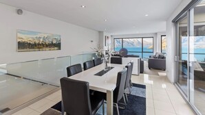 A modern dining area with a glass table and chairs, a large painting on the wall, and a view of the sea through large windows.