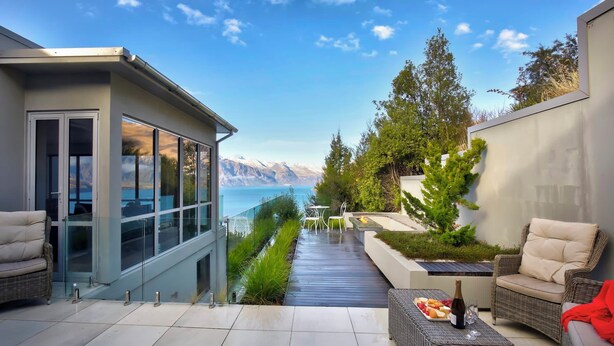 A modern outdoor patio with a view of mountains and the sea, furnished with wicker furniture and a glass door leading to a balcony.