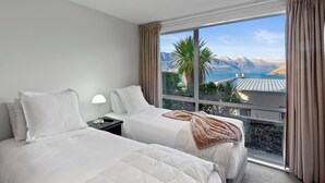 A hotel room with two beds, a nightstand, a lamp, and a large window with a view of mountains and a lake.