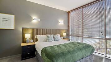 Une chambre moderne avec un grand lit, deux tables de chevet avec des lampes, une tête de lit avec des lattes verticales et une grande fenêtre avec des stores.