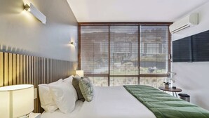 A modern hotel room with a bed, bedside table, lamp, and a view of the outdoors through large windows.