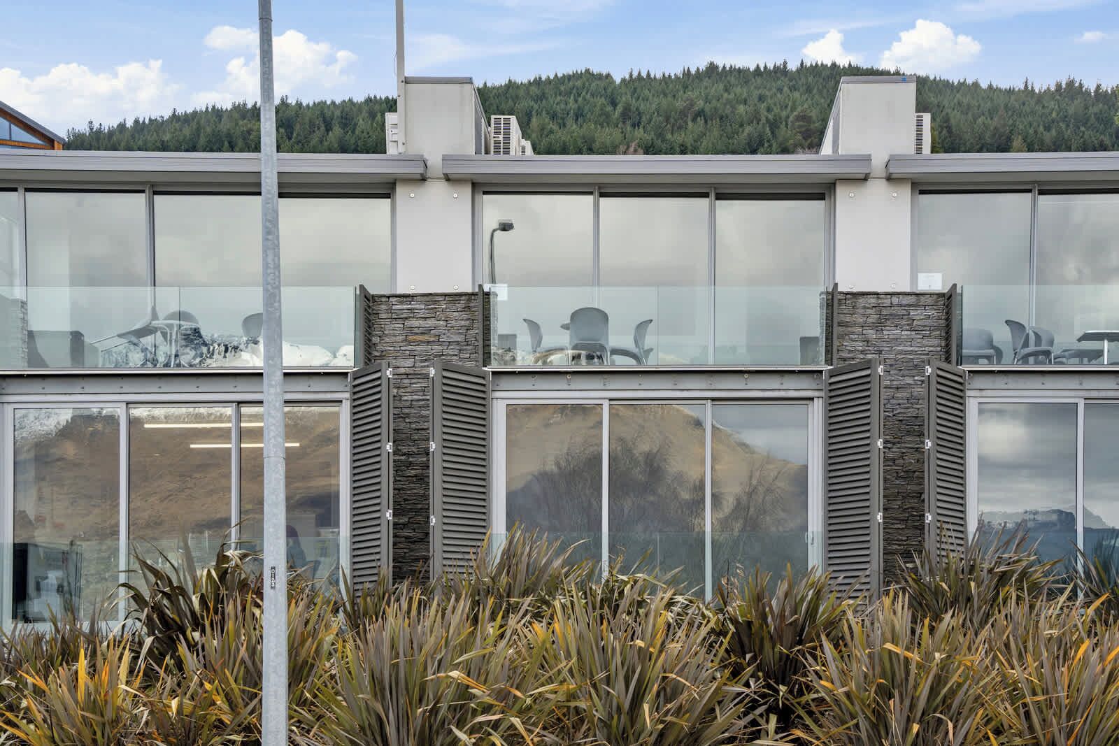Un bâtiment moderne doté de grandes fenêtres en verre, de piliers en pierre et offrant une vue sur une colline boisée.