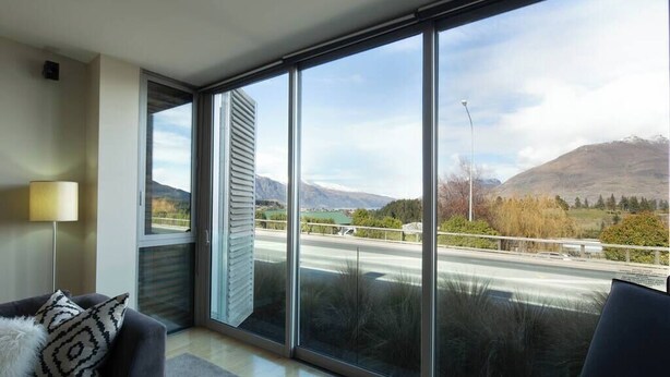 A modern living room with large windows, a sofa, a lamp, and a view of a highway and mountains. - Mountain View 2 Bed Apartment Pounamu 216 (Queenstown)