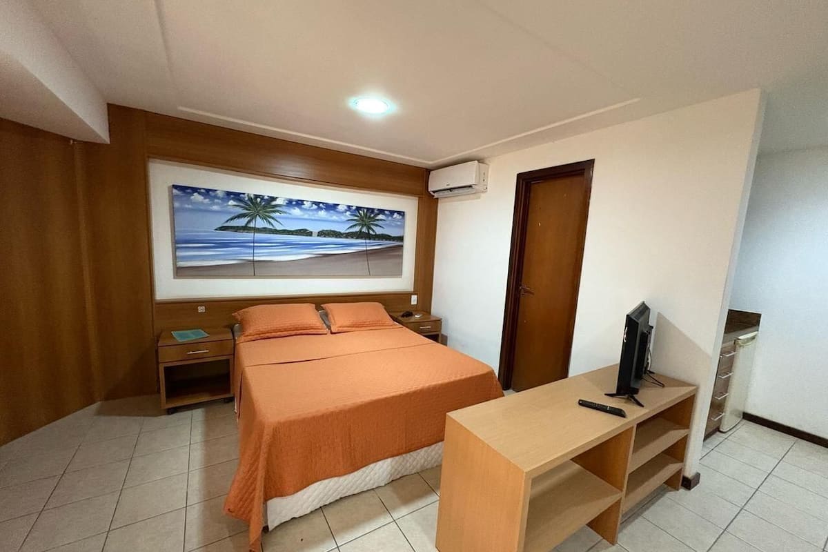 Un dormitorio con cama, escritorio, televisor, aire acondicionado y un cuadro en la pared.