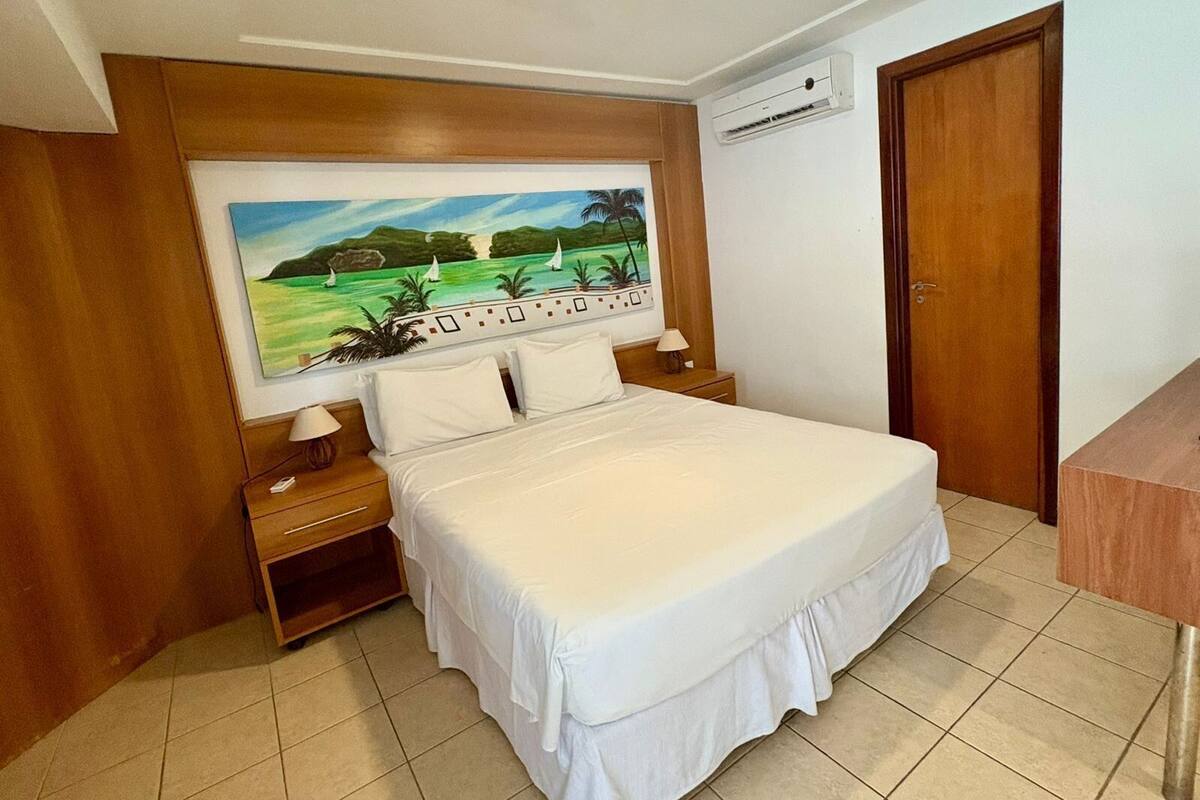 Un dormitorio con una cama grande, paneles de madera, un cuadro de un paisaje, un equipo de aire acondicionado y una puerta de madera.