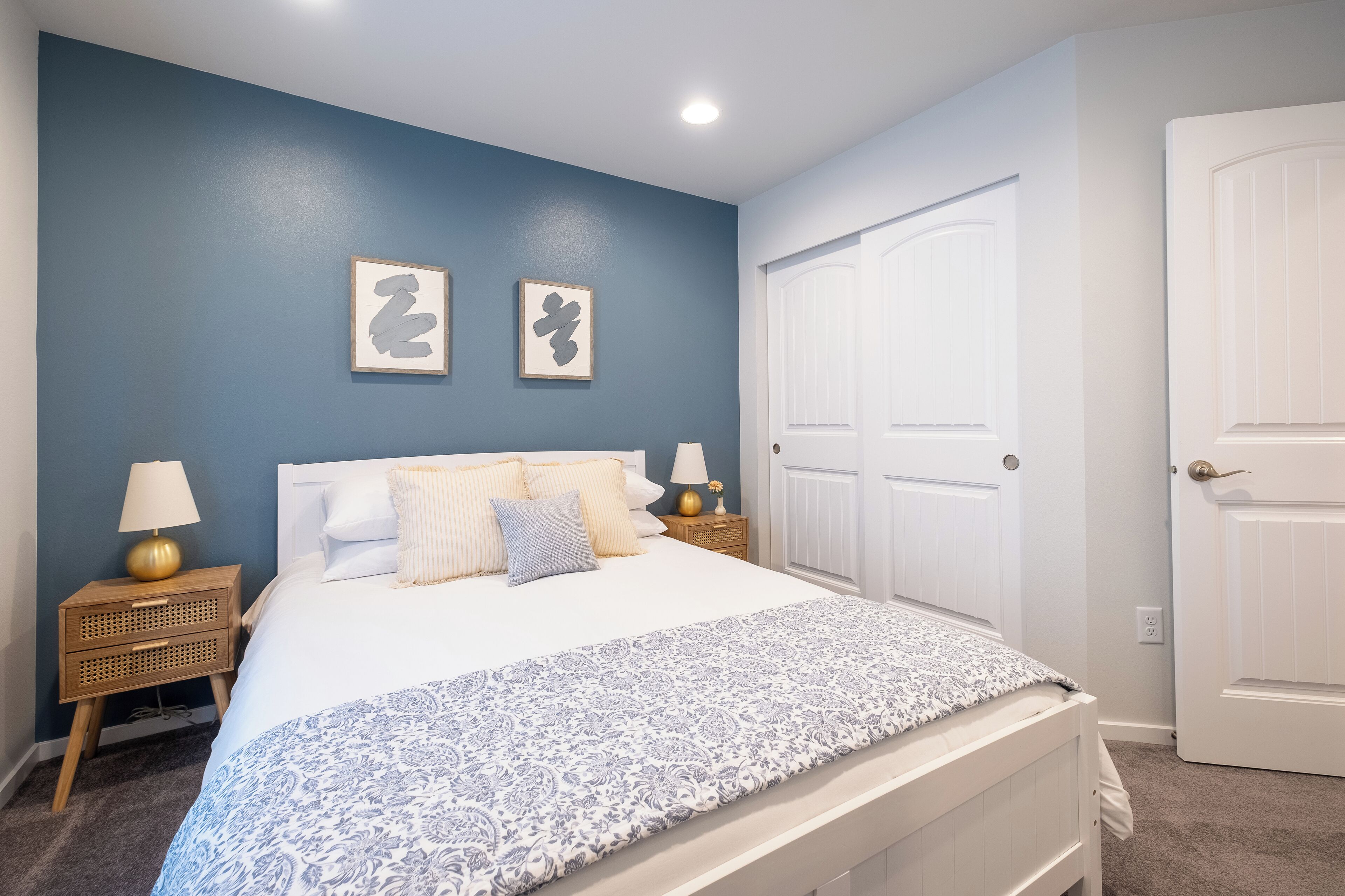 Une chambre à coucher avec un lit, deux tables de chevet avec des lampes, une porte et deux tableaux encadrés accrochés au mur.