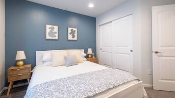 Une chambre à coucher avec un lit, deux tables de chevet avec des lampes, une porte et deux tableaux encadrés accrochés au mur.