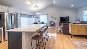 A modern kitchen with stainless steel appliances, a central island with bar stools, and a dining area with a sofa and TV. - Water & Mountain Views: Escape to Whidbey Island! (Freeland)