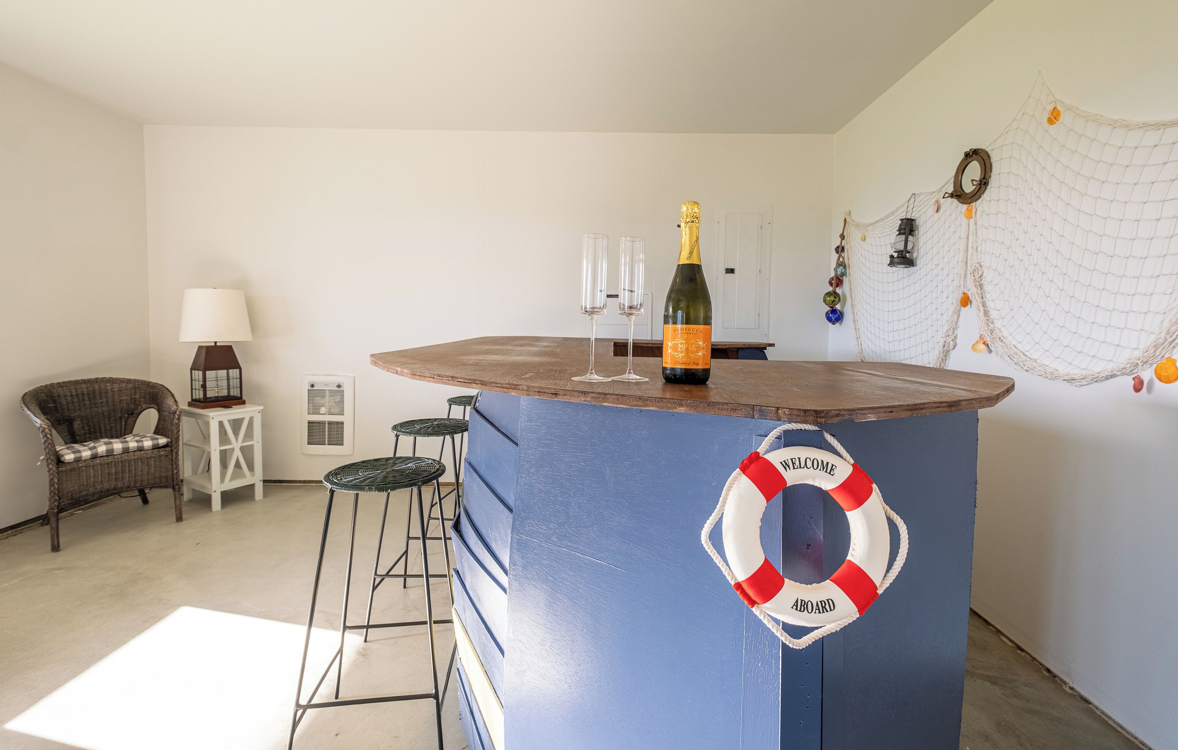 Zona de bar con un adorno de boya salvavidas, una botella de champán y dos copas sobre una barra de madera.